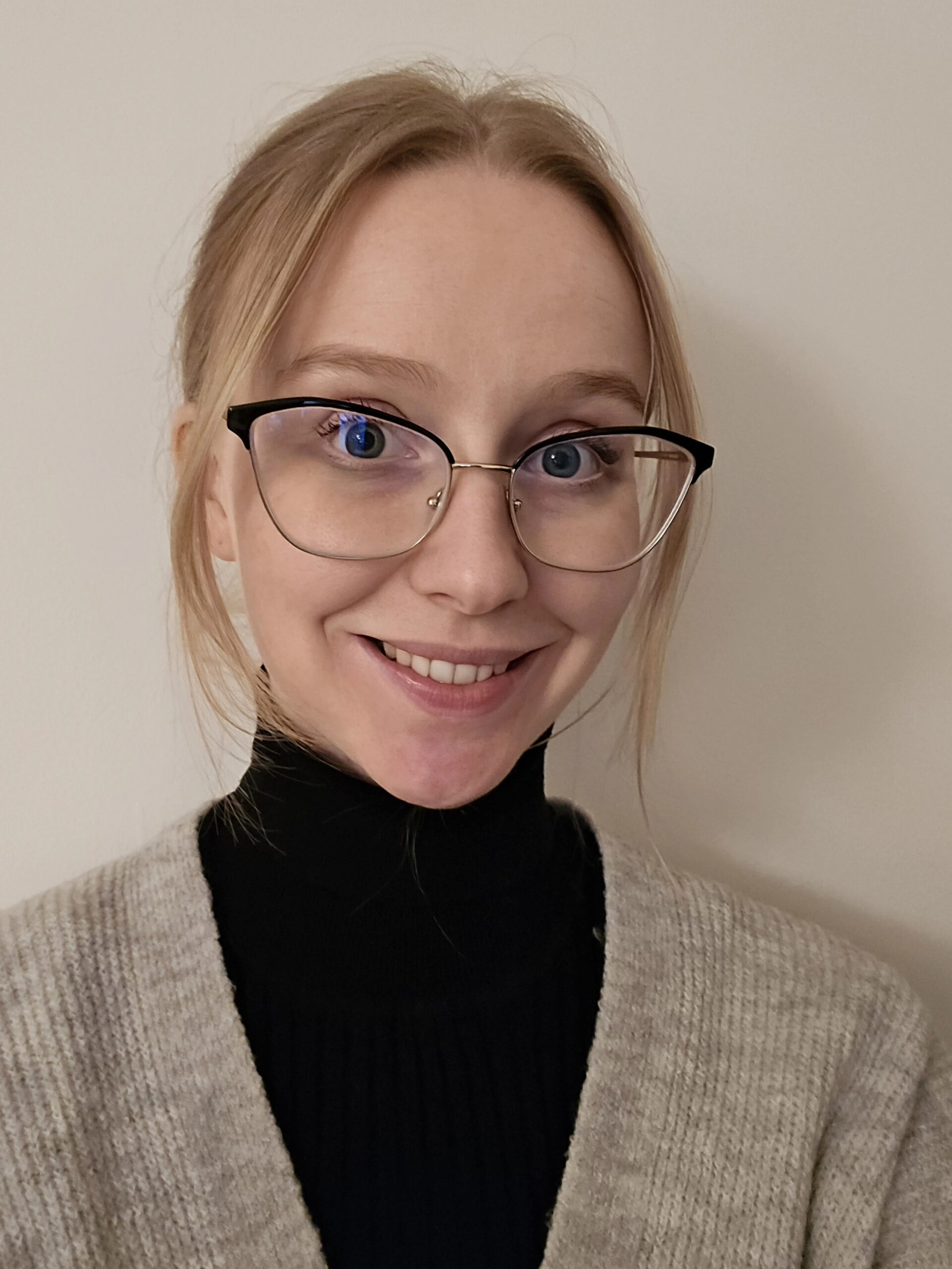 Picture of smiling young woman with glasses and blonde hair. Wearing a black turtle neck top and Taupe cardigan.